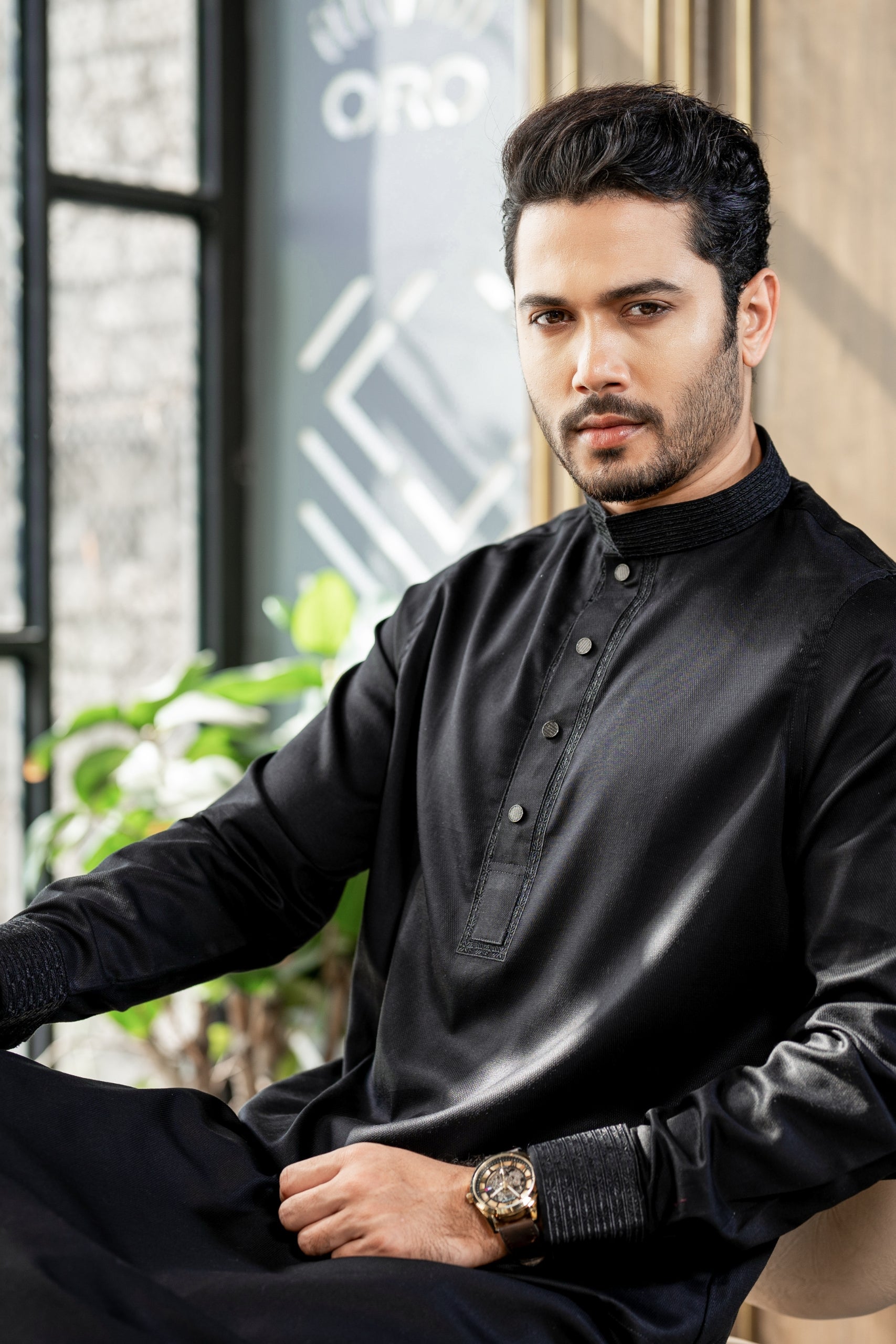 Man wearing a black traditional outfit sitting indoors with a blurred background
