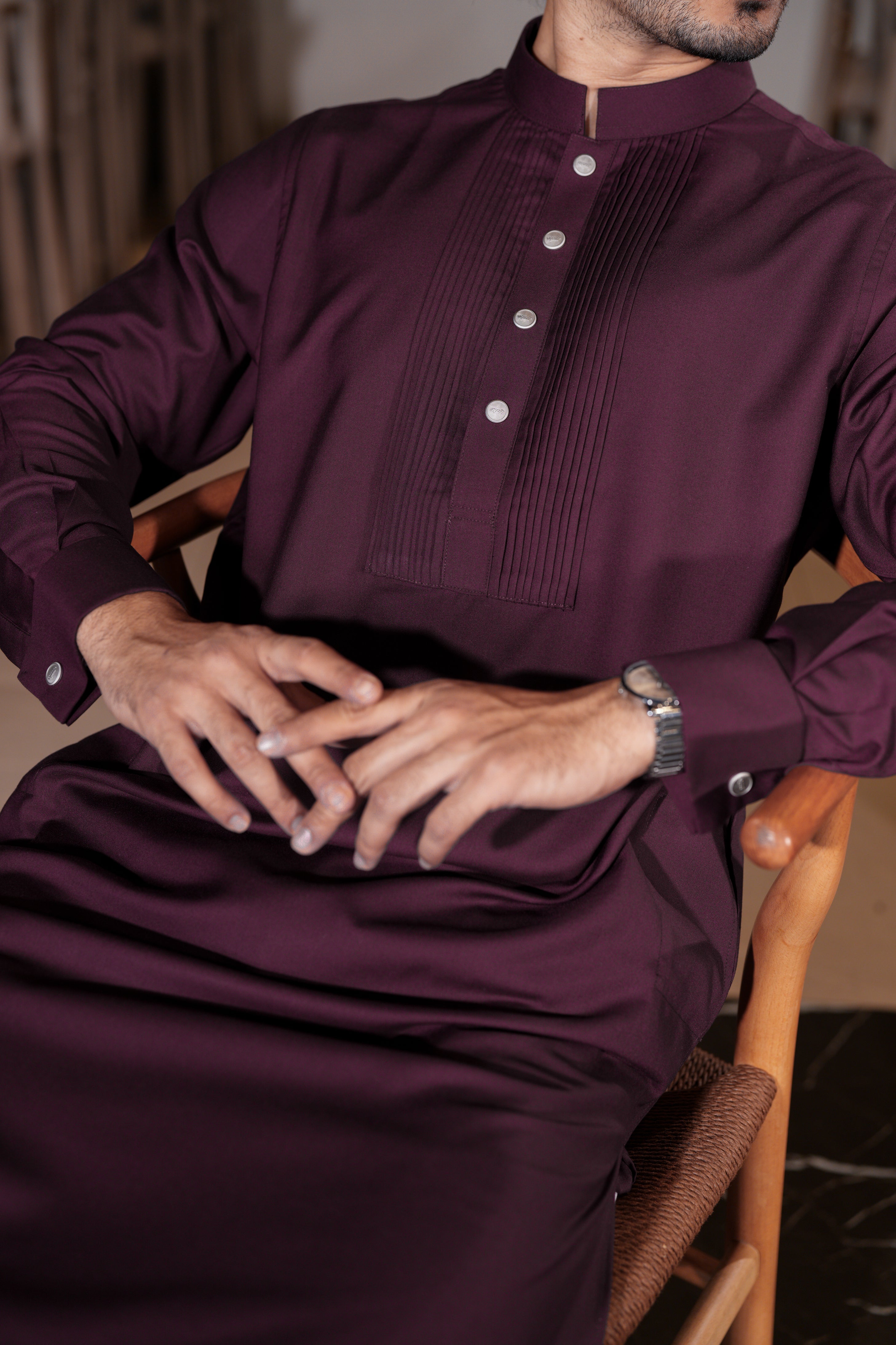 Man wearing a dark purple traditional outfit sitting on a wooden chair.