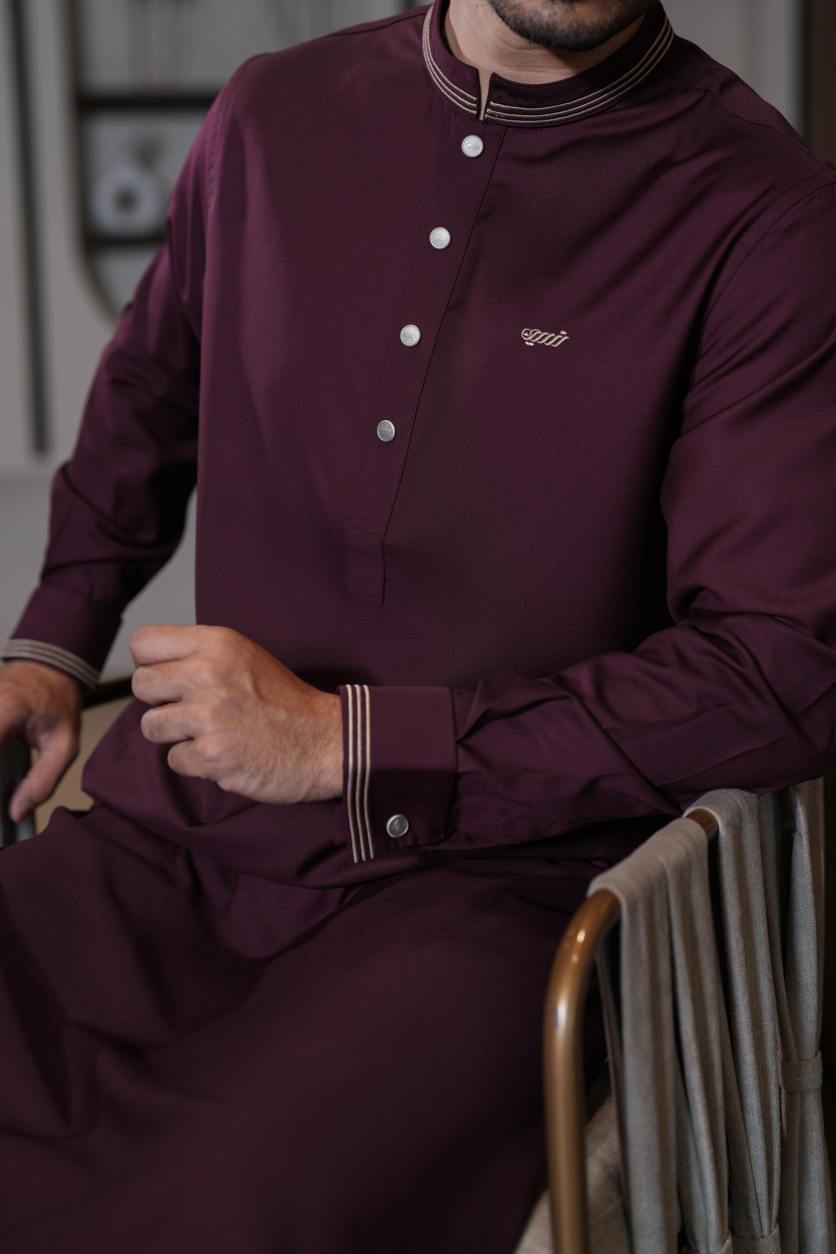 Man wearing a burgundy traditional outfit with buttons, sitting indoors.