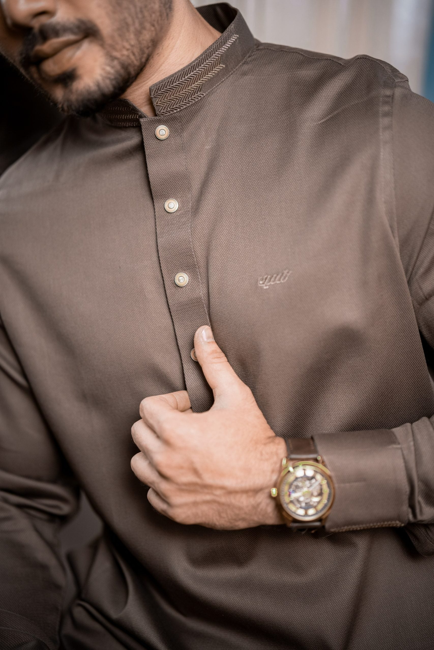 Man wearing a brown shirt with a brand logo, holding his shirt collar.