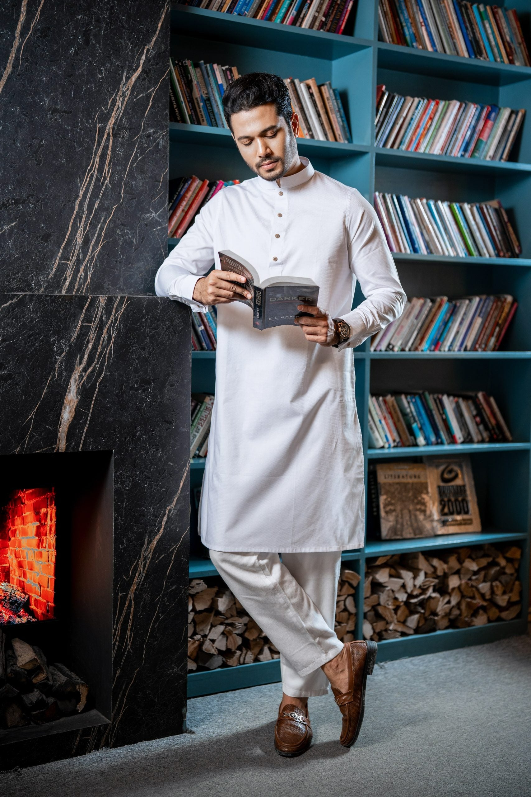 Man in a white kurta standing in front of a bookshelf with books and a fireplace.