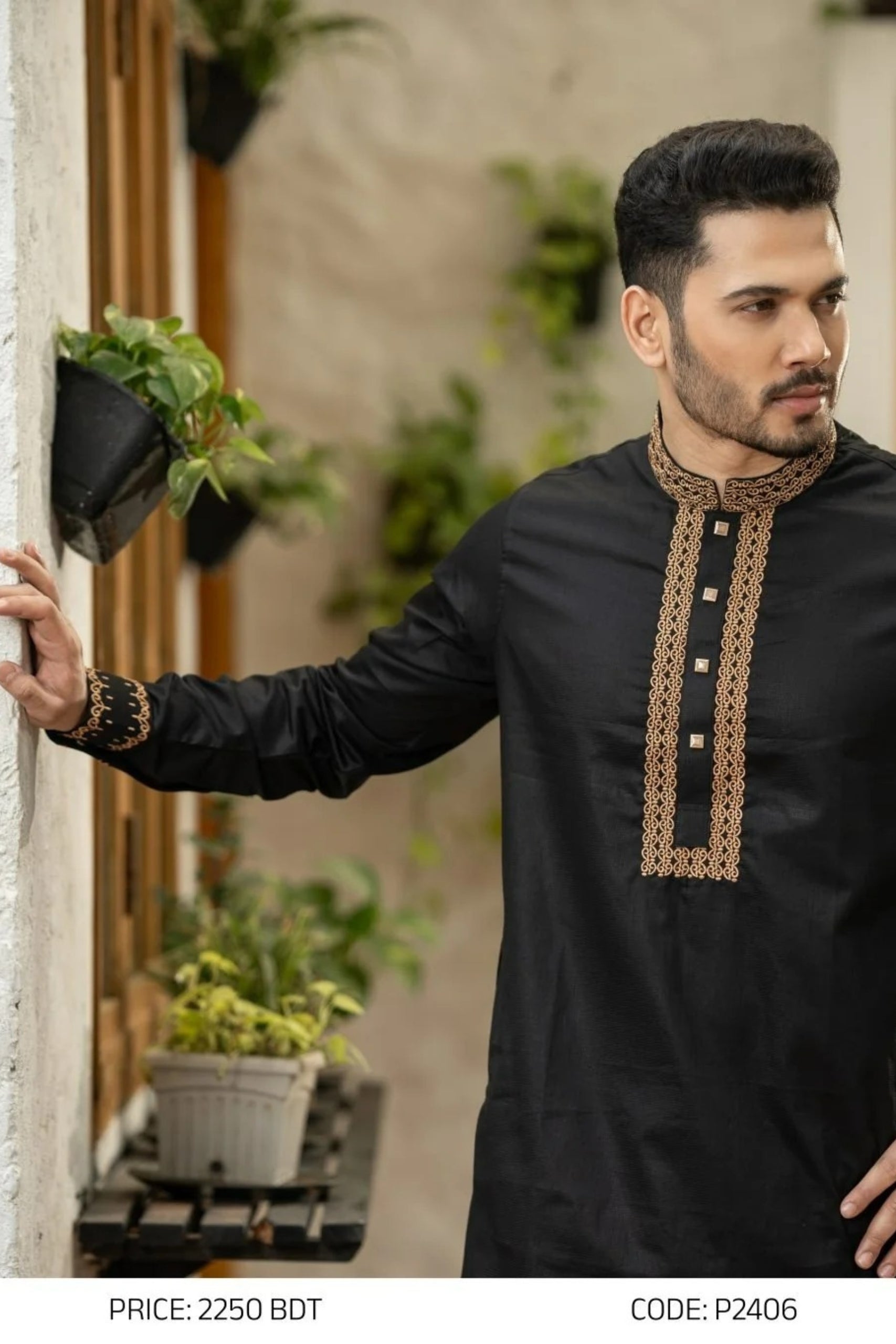 Man wearing a black kurta with intricate embroidery, standing in an outdoor setting with plants.