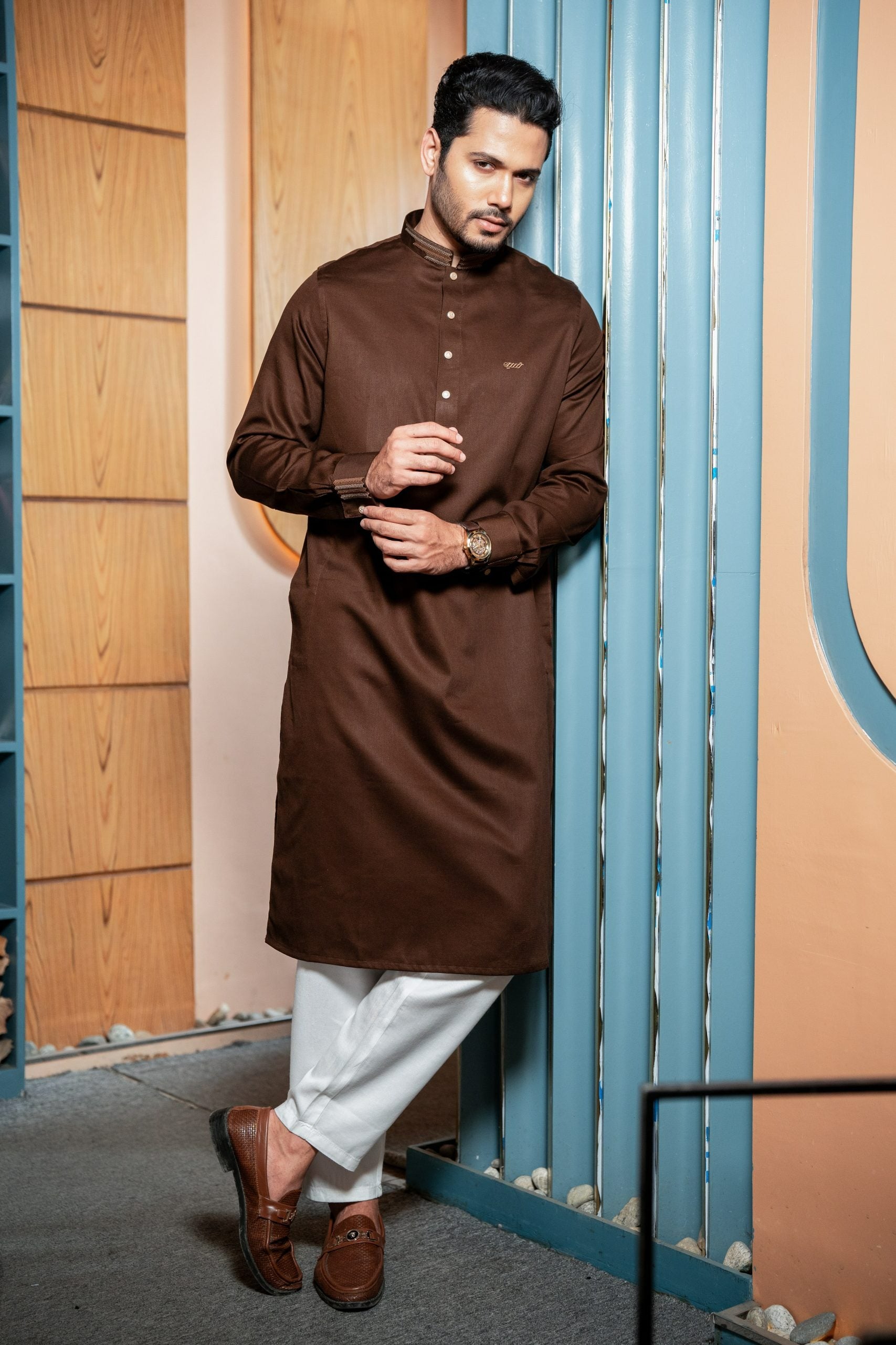 Man wearing a brown kurta with white pants standing against a colorful wall.